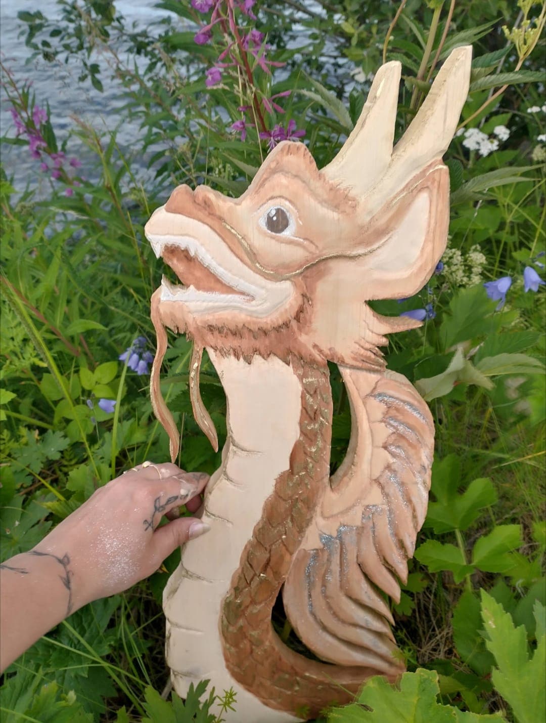 A hand holding a dragon carved from pale wood, upright among fireweed and harebells by the water